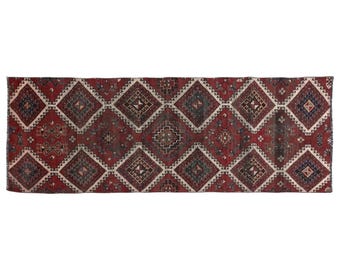 Puede incluir: Una alfombra larga y rectangular con un patrón de diamantes repetido. La alfombra presenta un fondo rojo intenso con diseños geométricos en tonos crema, azul marino y gris. El diseño de la alfombra es simétrico, con cada diamante que contiene detalles intrincados.