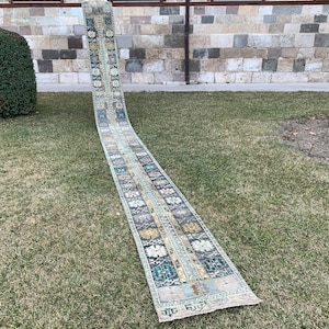 May include: A long, narrow rug with a traditional Persian design in shades of blue, beige, and yellow. The rug is laid out on a grassy lawn.