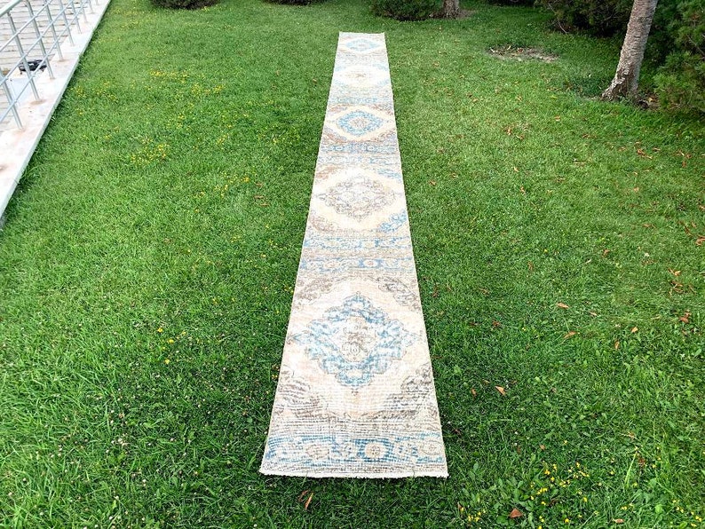 May include: A long, rectangular rug with a cream background and a blue and brown geometric pattern. The rug is laid out on a green grassy lawn.