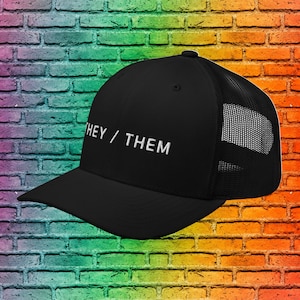May include: Black trucker hat with a white mesh back and the text "THEY/THEM" embroidered in white on the front.
