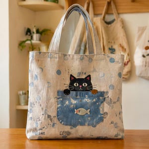 Könnte beinhalten: Eine helle Tragetasche mit blauem Tie-Dye-Punktemuster. Auf der Vorderseite befindet sich eine aufgesetzte Tasche aus blauem Denim, aus der ein gestickter schwarzer Kater mit weißen Augen und rosa Ohren schaut, sowie ein kleiner gestickter Fisch.
