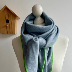 Può includere: Una sciarpa grigio chiaro con bordi verde brillante è drappeggiata su un manichino. La sciarpa è annodata al collo e cade in morbide pieghe. Una piccola casetta per uccelli in legno è visibile sullo sfondo.