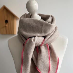 Puede incluir: Bufanda beige con ribete rosa brillante drapeada sobre un maniquí. La bufanda está anudada en el cuello y cuelga en suaves pliegues. En el fondo se ve un pequeño nido de pájaros de madera.