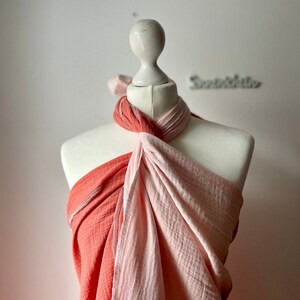 Può includere: Un'avvolgente in mussola corallo e rosa è drappeggiata su un manichino bianco. L'avvolgente è annodata al collo e le estremità pendono.
