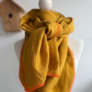 Puede incluir: Bufanda de color amarillo mostaza con ribete naranja, drapeada sobre un maniquí. La bufanda está hecha de una tela texturizada y está anudada en el cuello. El ribete naranja brillante proporciona un contraste llamativo con el amarillo.