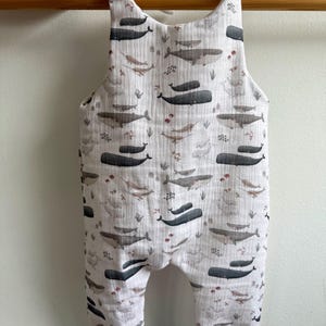 K&ouml;nnte beinhalten: Wei&szlig;er Baby-Strampler mit Walmuster. Der Strampler hat ein sich wiederholendes Muster aus grauen Walen, Meerespflanzen und anderen Meeresbewohnern. Die Tr&auml;ger sind an den Schultern gebunden. Die B&uuml;ndchen an den Kn&ouml;cheln sind grau.