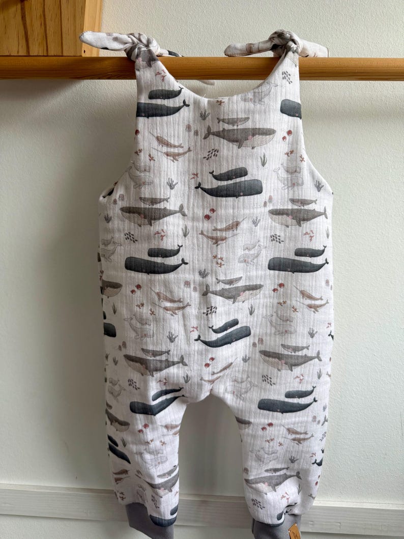 K&ouml;nnte beinhalten: Wei&szlig;er Baby-Strampler mit Walmuster. Der Strampler hat ein sich wiederholendes Muster aus grauen Walen, Meerespflanzen und anderen Meeresbewohnern. Die Tr&auml;ger sind an den Schultern gebunden. Die B&uuml;ndchen an den Kn&ouml;cheln sind grau.