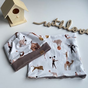 Puede incluir: Un conjunto de gorro y braga de cuello blancos con temática de circo. La tela presenta artistas de circo y animales, con un ribete marrón. El conjunto está diseñado para la comodidad y el estilo, ideal para un armario infantil.