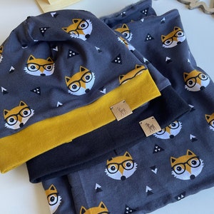 Puede incluir: Un conjunto de dos accesorios de temática de zorro gris y negro. La boina tiene un borde amarillo y un patrón de zorros con gafas y triángulos. La bufanda a juego tiene el mismo patrón. Ambos artículos tienen una pequeña etiqueta de cuero marrón con una silueta de ciervo.