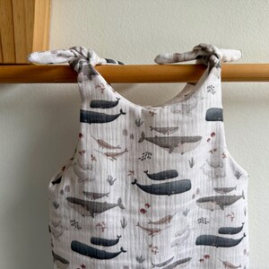 K&ouml;nnte beinhalten: Ein wei&szlig;er Baby-Strampler mit Walmuster. Der Strampler hat ein sich wiederholendes Muster aus grauen und schwarzen Walen, Narwalen und Seetang. Die Tr&auml;ger sind an den Schultern gebunden. Der Strampler h&auml;ngt an einer Holzstange.