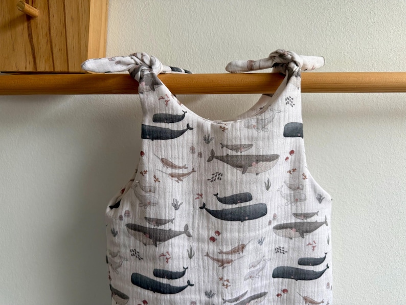 K&ouml;nnte beinhalten: Ein wei&szlig;er Baby-Strampler mit Walmuster. Der Strampler hat ein sich wiederholendes Muster aus grauen und schwarzen Walen, Narwalen und Seetang. Die Tr&auml;ger sind an den Schultern gebunden. Der Strampler h&auml;ngt an einer Holzstange.