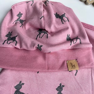 Puede incluir: Un gorro de punto rosa con un dobladillo y una etiqueta de cuero marrón con una silueta de ciervo. El gorro está cubierto de un patrón repetido de siluetas de ciervos negros.