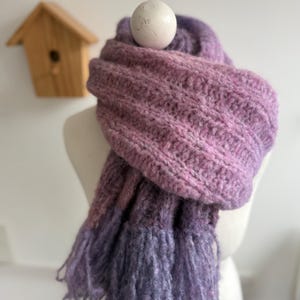 Könnte beinhalten: Handgestrickter Schal in Lila- und Rosatönen mit Fransen. Der Schal wird auf einer Schaufensterpuppe präsentiert, die seine Textur und den Farbverlauf zeigt. Im Hintergrund ist ein Vogelhaus aus Holz zu sehen.