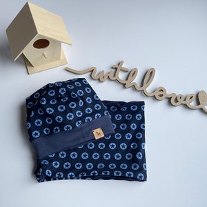 Puede incluir: Un gorro y una bufanda a juego de punto azul con un patrón de estrellas blancas. El gorro tiene un borde doblado y la bufanda está doblada por la mitad. Las palabras "with love" están escritas en un letrero de madera en el fondo.