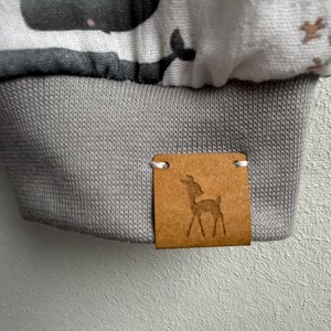 K&ouml;nnte beinhalten: Nahaufnahme eines hellgrauen Stoffes mit einem dunkelgrauen Walmuster. Der untere Rand hat eine graue Rippb&uuml;ndchen. Ein kleines braunes Lederetikett mit einer Hirschsilhouette ist angebracht.