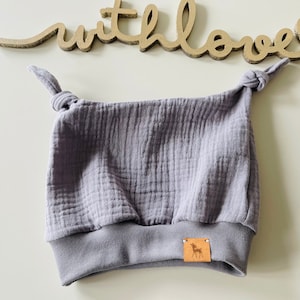 Peut inclure: Un bonnet gris pour bébé avec des détails noués sur le dessus et un bord assorti. Le chapeau est en tissu texturé et comporte une petite étiquette en cuir marron avec une silhouette de cerf. Le mot "with love" est en lettres dorées en arrière-plan.