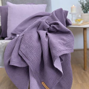 Könnte beinhalten: Eine tiefviolette Musselindecke mit knittriger Textur und einem kleinen rechteckigen, braunen lederartigen Etikett an der Ecke. Der Stoff ist über einen Stuhl drapiert und zeigt sein weiches, geschichtetes Aussehen und die einheitliche Farbe.