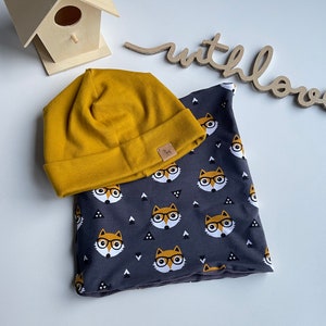 Puede incluir: Un gorro de punto amarillo mostaza y un cuello de tela gris con un patrón repetido de zorros con gafas. Los zorros son naranja y blanco con ojos negros y gafas negras.
