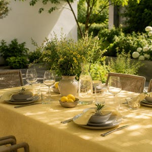 Op de afbeelding: Een prachtig gedekte eettafel buiten met een gele tafelkleed. De tafel is gedekt met borden, kommen, bestek en wijnglazen. Een vaas met gele bloemen en citroenen staat in het midden. Rotan stoelen omringen de tafel.