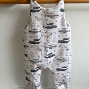 K&ouml;nnte beinhalten: Wei&szlig;er Baby-Strampler mit Walmuster in Grau- und Braunt&ouml;nen. Der Strampler hat graue B&uuml;ndchen und Tr&auml;ger zum Binden. Der Stoff ist ein weiches, leichtes Material.