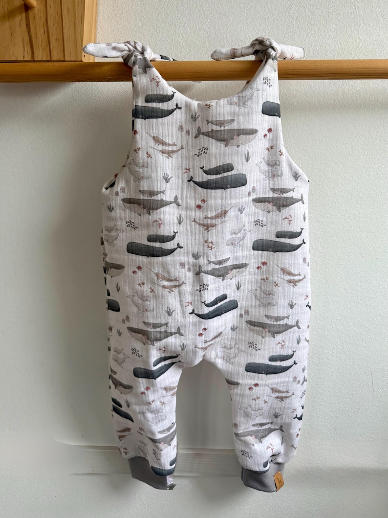 K&ouml;nnte beinhalten: Wei&szlig;er Baby-Strampler mit Walmuster in Grau- und Braunt&ouml;nen. Der Strampler hat graue B&uuml;ndchen und Tr&auml;ger zum Binden. Der Stoff ist ein weiches, leichtes Material.