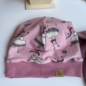 Puede incluir: Gorro de punto rosa con un patrón de chicas con vestidos blancos y coronas. El gorro tiene un puño abatible y una pequeña etiqueta de logo de ciervo marrón.