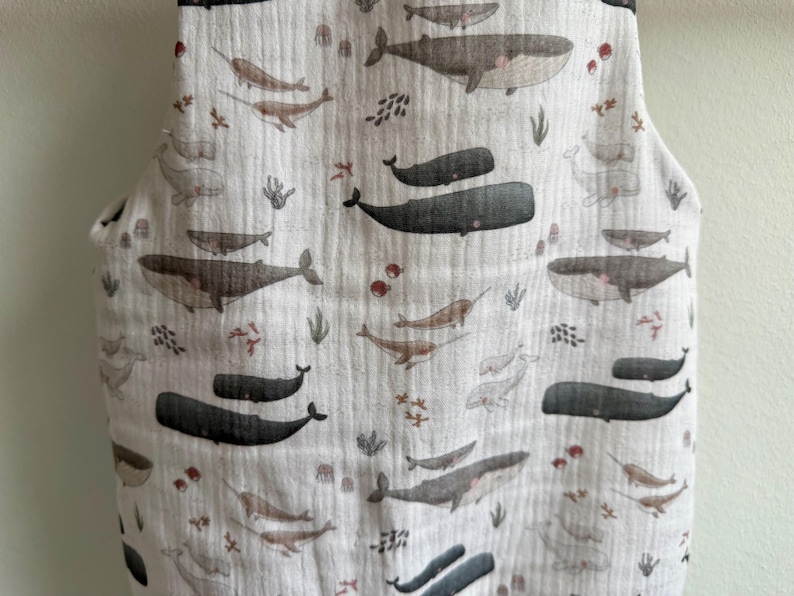 K&ouml;nnte beinhalten: Ein wei&szlig;er Stoff mit einem sich wiederholenden Muster aus Walen, Narwalen und Wasserpflanzen in Grau-, Braun- und Rott&ouml;nen. Der Stoff hat ein strukturiertes, geknittertes Aussehen. Das Design zeigt Wale und Narwale in verschiedenen Gr&ouml;&szlig;en.