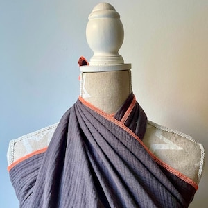 Può includere: Un capo di tessuto grigio drappeggiato con finiture arancioni è esposto su un manichino. Il tessuto è avvolto attorno al collo e alle spalle del manichino, creando un effetto a strati. Le finiture arancioni aggiungono un tocco di colore.