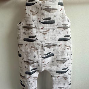 K&ouml;nnte beinhalten: Ein wei&szlig;er Baby-Strampler mit einem Walmuster in Grau- und Braunt&ouml;nen. Der Strampler hat graue B&uuml;ndchen und Bindetr&auml;ger an den Schultern. Der Stoff ist ein weiches, strukturiertes Material.