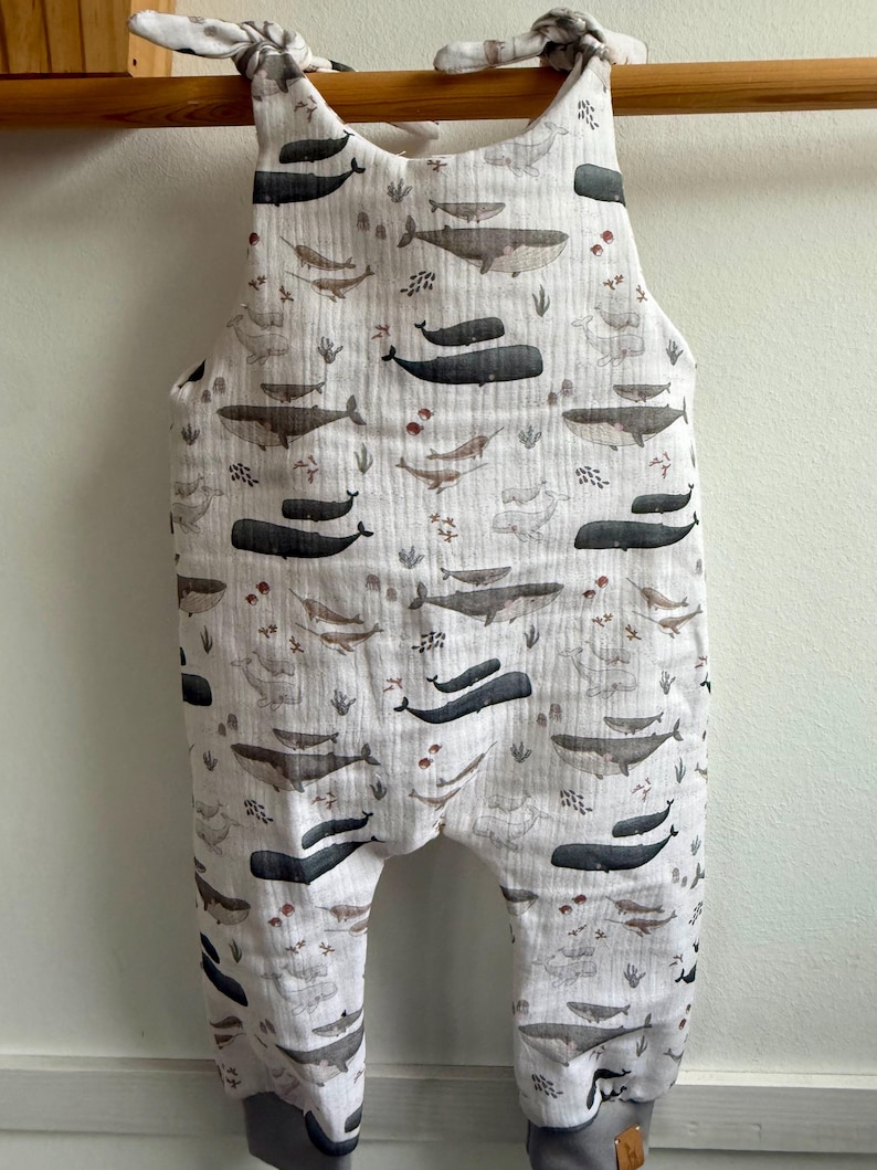 K&ouml;nnte beinhalten: Ein wei&szlig;er Baby-Strampler mit einem Walmuster in Grau- und Braunt&ouml;nen. Der Strampler hat graue B&uuml;ndchen und Bindetr&auml;ger an den Schultern. Der Stoff ist ein weiches, strukturiertes Material.