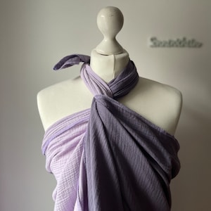 Può includere: Un'avvolgente in mussola di colore viola e lavanda è legata attorno a un manichino bianco. L'avvolgente è realizzata in un tessuto morbido e traspirante, ideale per il babywearing.
