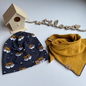 Puede incluir: Un gorro gris oscuro con un estampado de zorros con gafas, combinado con una bufanda de color amarillo mostaza. Una pequeña casa para pájaros de madera y un letrero de madera con la inscripción "with love" también están en la imagen.