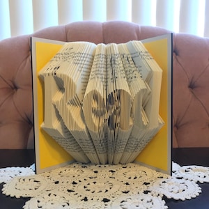 May include: A book art sculpture of the word "ROCK" made by folding the pages of a book. The sculpture is displayed on a white doily on a black surface.