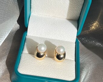 May include: A pair of gold pearl earrings in a teal jewelry box. The earrings are made of gold and have a single pearl on each earring.