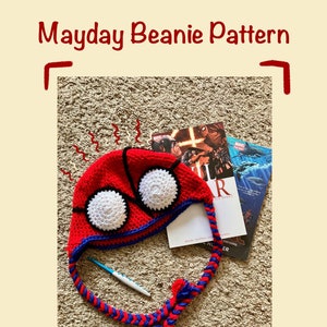May include: A red and blue crocheted beanie hat with white eyes. The hat has a long, braided cord. The hat is lying on a brown carpet with a book and a crochet hook.