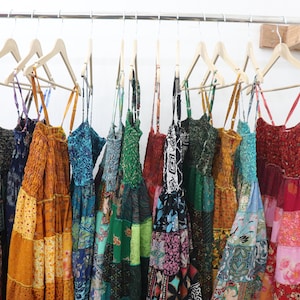 Könnte beinhalten: Ein Kleiderständer mit bunten Patchwork-Kleidern mit dünnen Trägern, die an Holzbügeln hängen. Die Kleider sind aus verschiedenen Stoffen und Mustern gefertigt, darunter Blumen-, Paisley- und geometrische Muster.