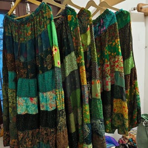 Könnte beinhalten: Mehrere lange Patchwork-Röcke hängen an Holzbügeln. Die Röcke weisen eine Vielzahl von grünen, türkisfarbenen und braunen Stoffen mit floralen und geometrischen Mustern auf. Die Röcke werden vor einem neutralen Hintergrund präsentiert.