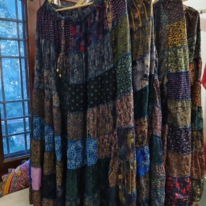 Può includere: Tre lunghe gonne di patchwork colorate appese a grucce di legno. Le gonne sono realizzate con diversi tessuti e motivi, tra cui disegni floreali, paisley e geometrici. Le gonne sono tutte di lunghezze diverse, con la gonna più lunga che arriva a terra.