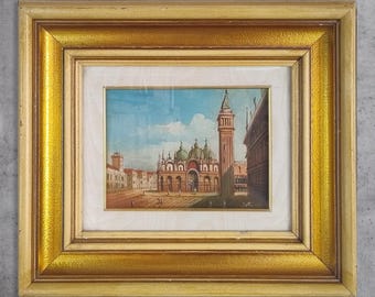 Puede incluir: Pintura enmarcada de un paisaje urbano europeo. La obra de arte representa una plaza con edificios, una torre alta y una estructura con cúpula. El marco es de color dorado con un acabado texturizado, y la pintura está colocada detrás de un passe-partout blanco.