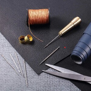 May include: A set of leatherworking tools including a spool of copper thread, two thimbles, a leather awl, four needles, and a pair of scissors.