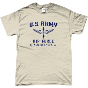 May include: A light tan t-shirt with the text "U.S. ARMY AIR FORCE MIAMI BEACH FLA." in navy blue. The design includes a propeller and wing graphic. The shirt is a crew neck style.