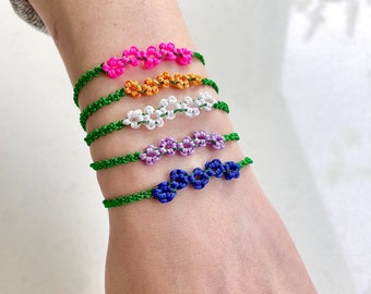May include: Five colorful friendship bracelets with green string and flower-shaped knots. The bracelets are stacked on a person's wrist.