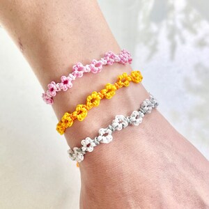 Puede incluir: Tres pulseras de la amistad con nudos en forma de flor. Las pulseras son rosa, amarilla y blanca.