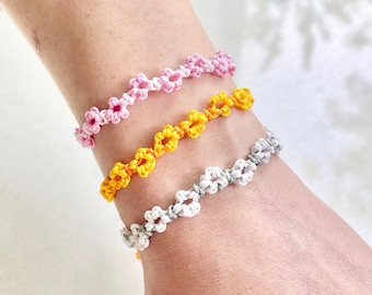 Puede incluir: Tres pulseras de la amistad con nudos en forma de flor. Las pulseras son rosa, amarilla y blanca.