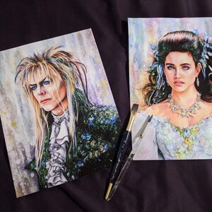 May include: Two watercolor paintings of characters from the movie Labyrinth. The painting on the left is of Jareth, the Goblin King, with long blond hair and a green and black outfit. The painting on the right is of Sarah Williams, with long brown hair and a white dress. Both paintings have a sparkly, glittery background.