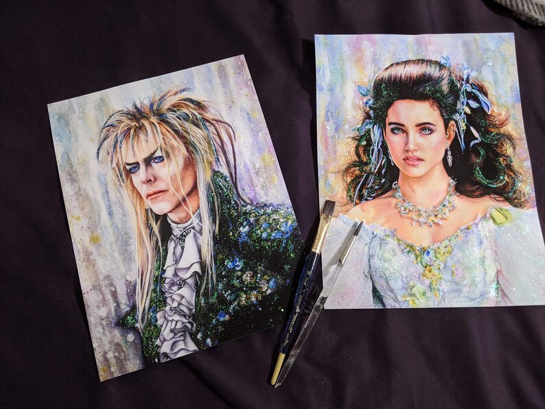 May include: Two watercolor paintings of characters from the movie Labyrinth. The painting on the left is of Jareth, the Goblin King, with long blond hair and a green and black outfit. The painting on the right is of Sarah Williams, with long brown hair and a white dress. Both paintings have a sparkly, glittery background.