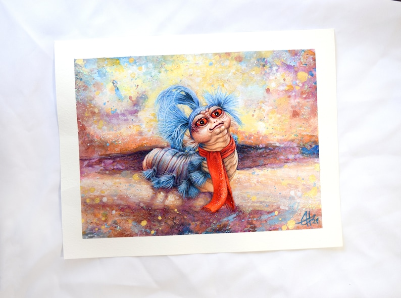 Puede incluir: Una pintura de acuarela de una criatura vermiforme azul y roja con una peluca azul y una bufanda roja. La criatura est&aacute; sentada sobre un fondo marr&oacute;n y amarillo con salpicaduras de pintura blanca y azul.