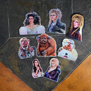 Puede incluir: Una colección de pegatinas troqueladas con personajes de la película Laberinto. Las pegatinas representan varios personajes, incluyendo a Jareth, Sarah y otras criaturas fantásticas. Las pegatinas tienen un borde brillante y están dispuestas sobre una superficie oscura y texturizada.