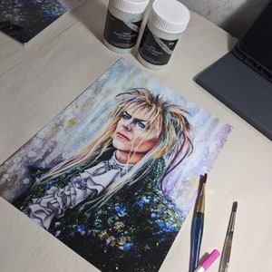 May include: A watercolor painting of a man with long, white hair and a black and green outfit. The painting is on a white background and has a sparkly, iridescent texture. There are three paintbrushes and two jars of iridescent glitter on the table.