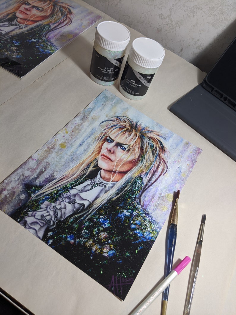 May include: A watercolor painting of a man with long, white hair and a black and green outfit. The painting is on a white background and has a sparkly, iridescent texture. There are three paintbrushes and two jars of iridescent glitter on the table.
